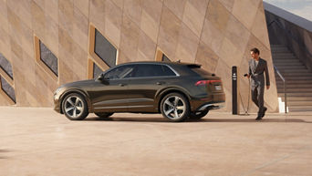Seitenaufnahme des Audi Q8 SUV TFSI e, der an einer Ladesäule parkt. Ein Mann im Anzug möchte gerade das Ladekabel anschließen.