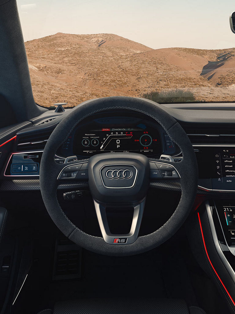 Blick auf das Cockpit eines Audi RS Q8 SUV mit Lenkrad, digitalen Anzeigen und Touchscreens für Klima und Fahrmodi und roter Ambientebeleuchtung.