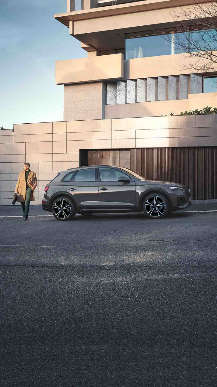 Vista lateral de un Audi Q5 color gris oscuro frente a un edificio, mientras un hombre con gabardina café pasa caminando.