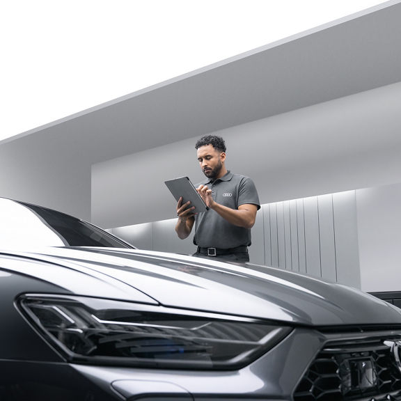 A male Audi Service representative using a tablet computer while standing next to a dark grey Audi.