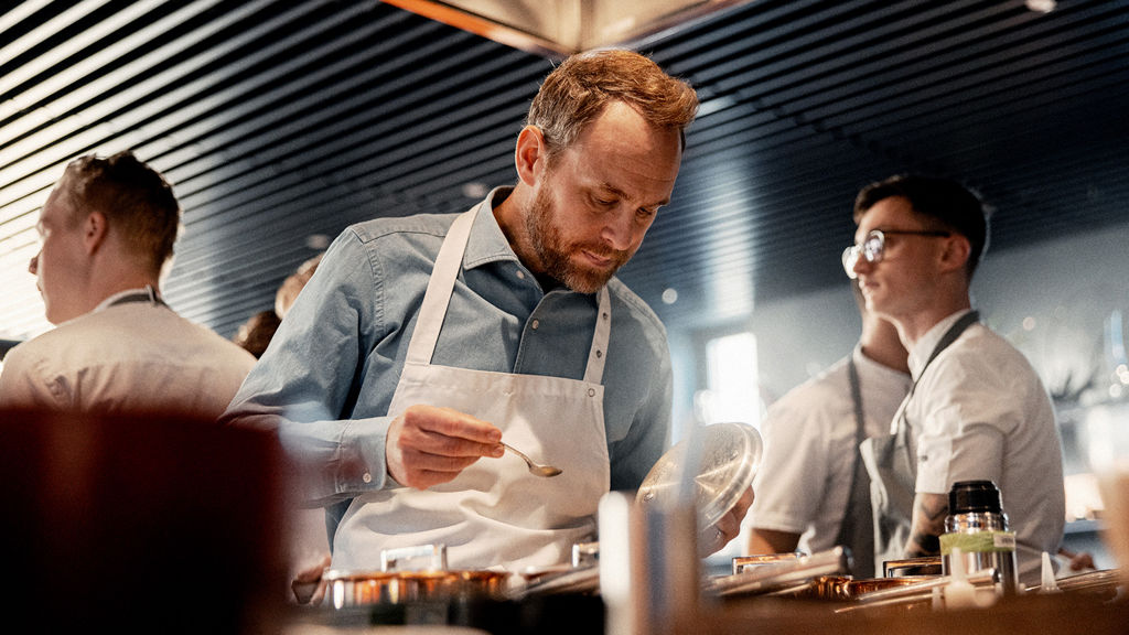 Björn Franzén visas framifrån hållande i ett grytlock samtidigt som han tittar ned i en gryta, en matsal men människor i bakgrunden.