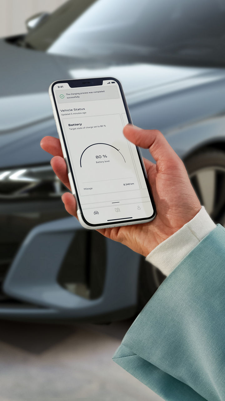 Kvinnas hand som håller i en telefon som visar myAudi-appen, bakom telefonen syns delar av en silvrig bil