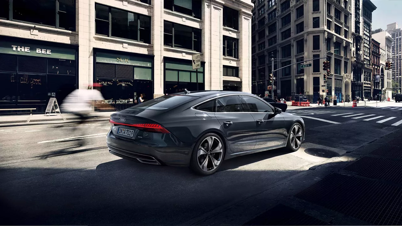 Ein dunkelgrauer Audi A7 Sportback fährt durch eine Straße in der Stadt, im Hintergrund hohe Gebäude und Passanten.