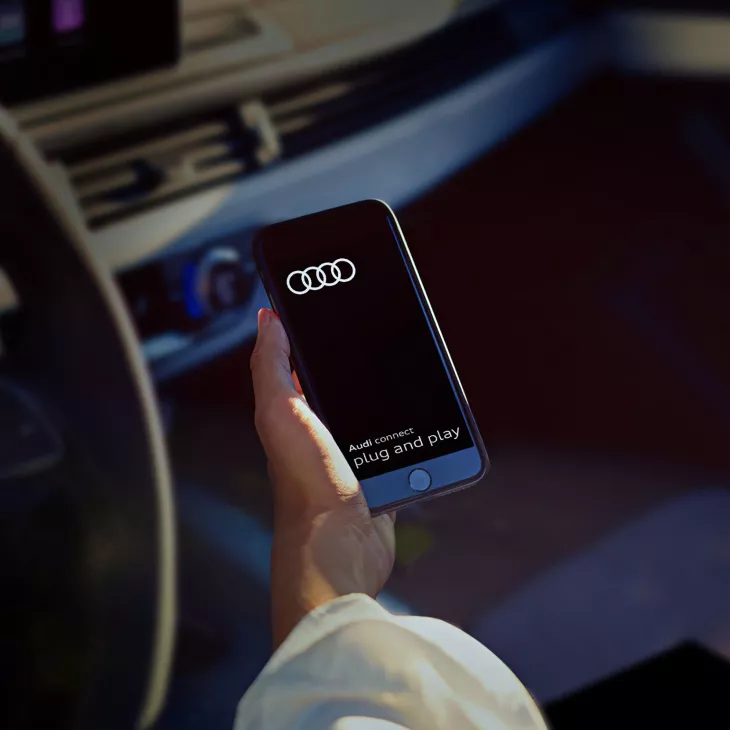 A person in an Audi vehicle, their hand holding a mobile phone, Audi Connect visible on its screen