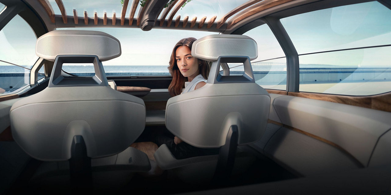 Interior view of a modern vehicle with wooden accents, a woman back at us in the passenger seat and a view of the ocean