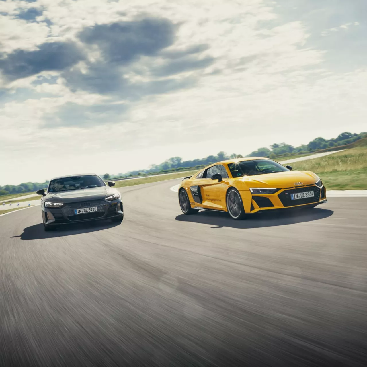 Two Audi vehicles racing on a track, one yellow and one black, under a cloudy sky