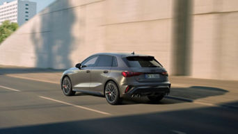 A silver Audi vehicle driving along a sunlit road beside a concrete wall