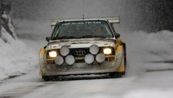 An Audi rally car racing through snow with headlights on