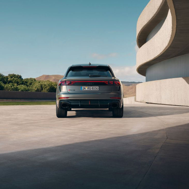 A silver Audi vehicle parked in front of a modern circular building with clear blue sky and distant mountains