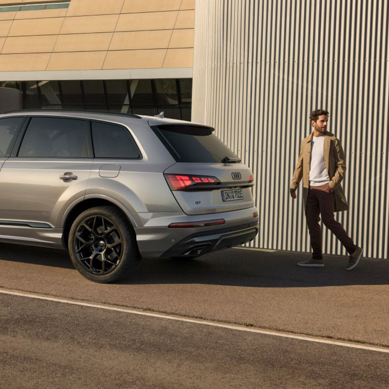 A person walking past a parked silver Audi Q7 vehicle near a corrugated metal wall