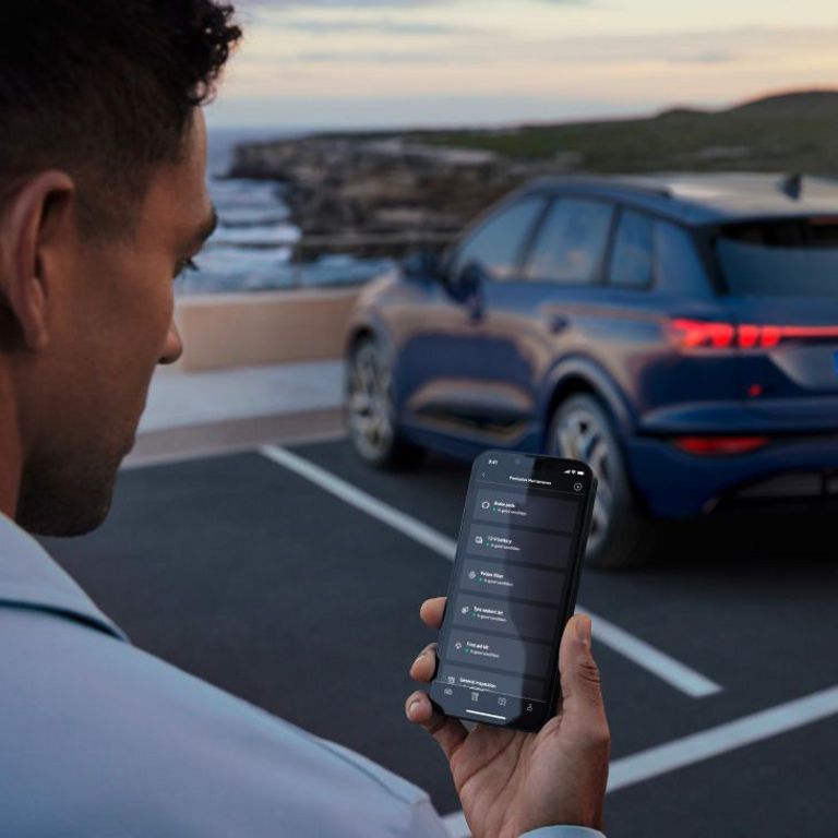 A person holding a smartphone with an app interface, standing near a parked Audi vehicle by the seaside