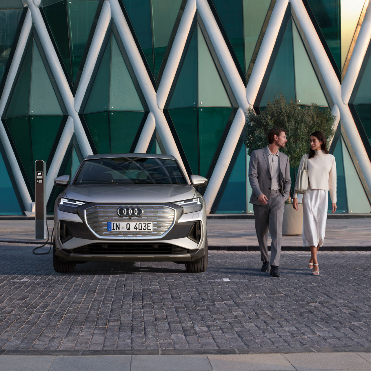 A young man and woman walking to the right of a parked Audi vehicle on charge at a charge station