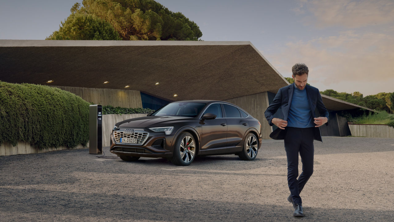 A man walking towards us with a parked black Audi electric vehicle on charge in the background 