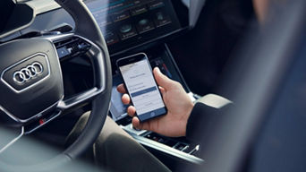 A person sat in the driver seat of an Audi, mobile phone in right hand displaying the myAudi app