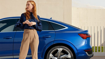 A young woman standing looking to her right standing in front of a parked blue Audi electric vehicle