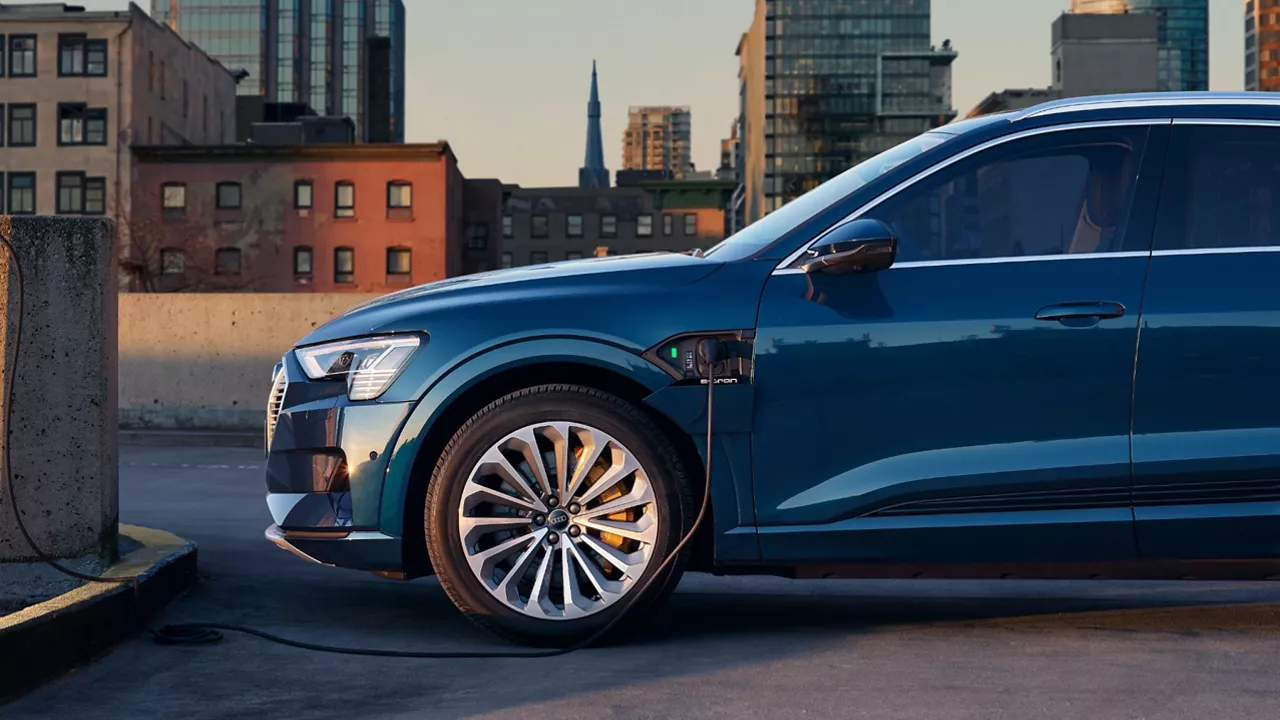 A parked blue Audi electric vehicle plugged in an on charge with a cityscape in the background