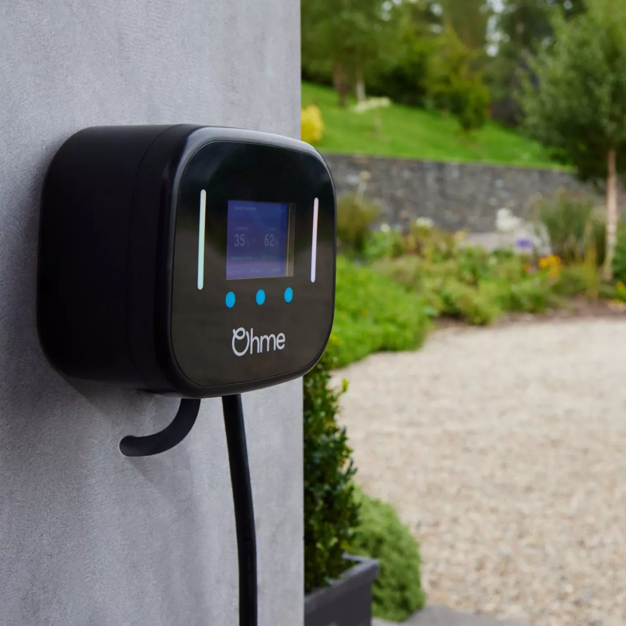 An Ohme electric vehicle home charger mounted to a house wall, gardens in the background