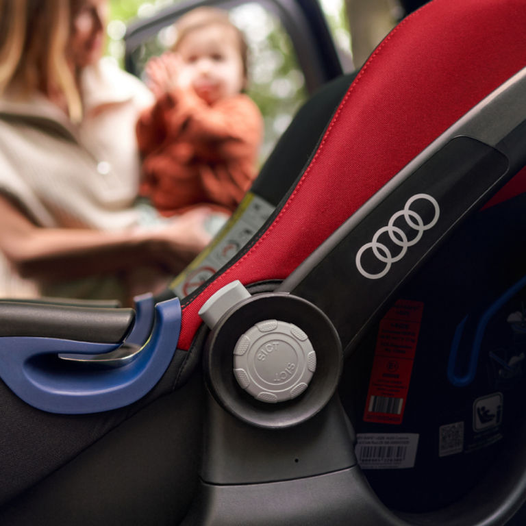 The interior of an Audi vehicle, focus on a Audi baby seat, mother and baby out of focus in distance