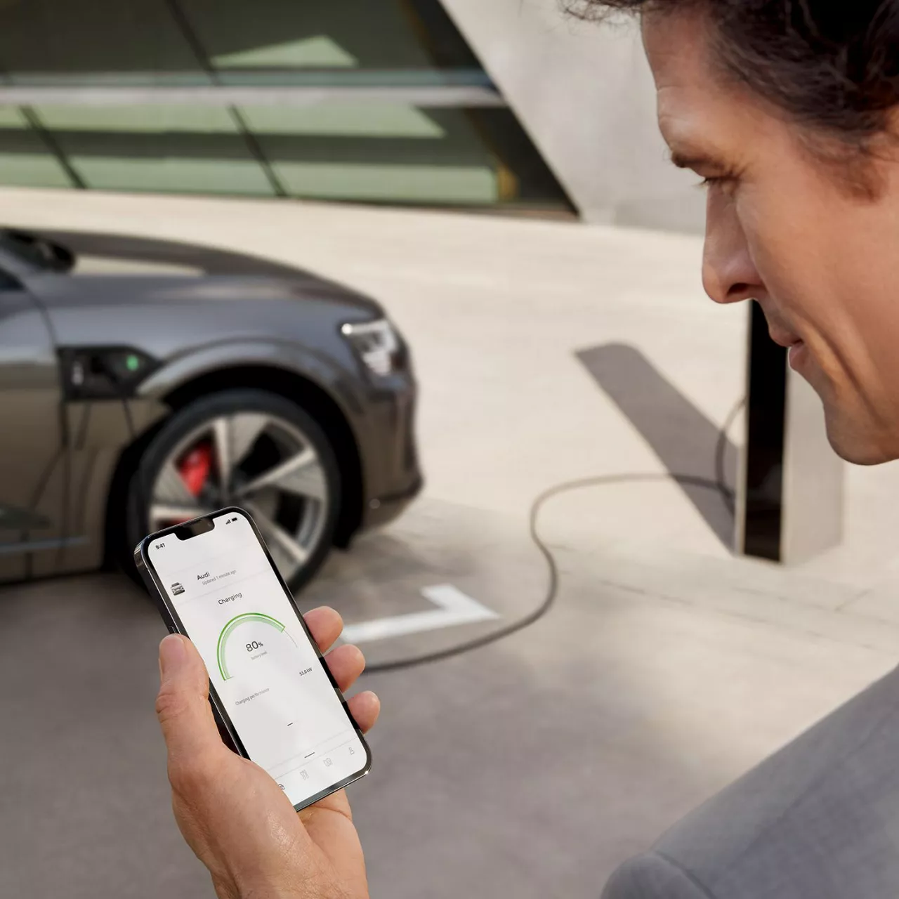 A man with his phone in his hand, the myAudi app on screen, Audi vehicle on charge in the background