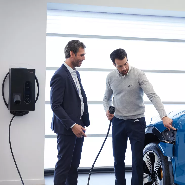 An Audi specialist, charge cable in hand, beside a customer pointing to the charge port on an Audi