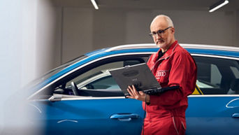 An Audi technician stood beside a parked Audi vehicle and using a laptop 