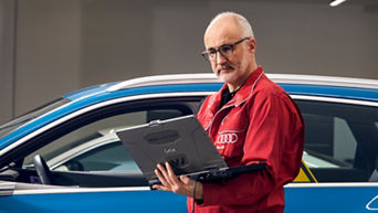 An Audi technician stood beside a parked Audi vehicle and using a laptop 