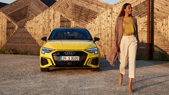 A young woman walking towards us to the right with a parked yellow Audi vehicle in the background