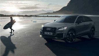 A beach in the late afternoon with a surfer walking on the left & a parked Audi vehicle on the right