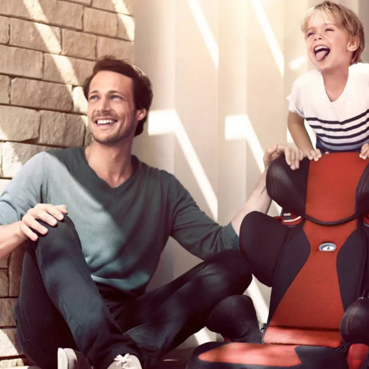 Two individuals sitting on a red chair beside a brick wall, one adult and one child