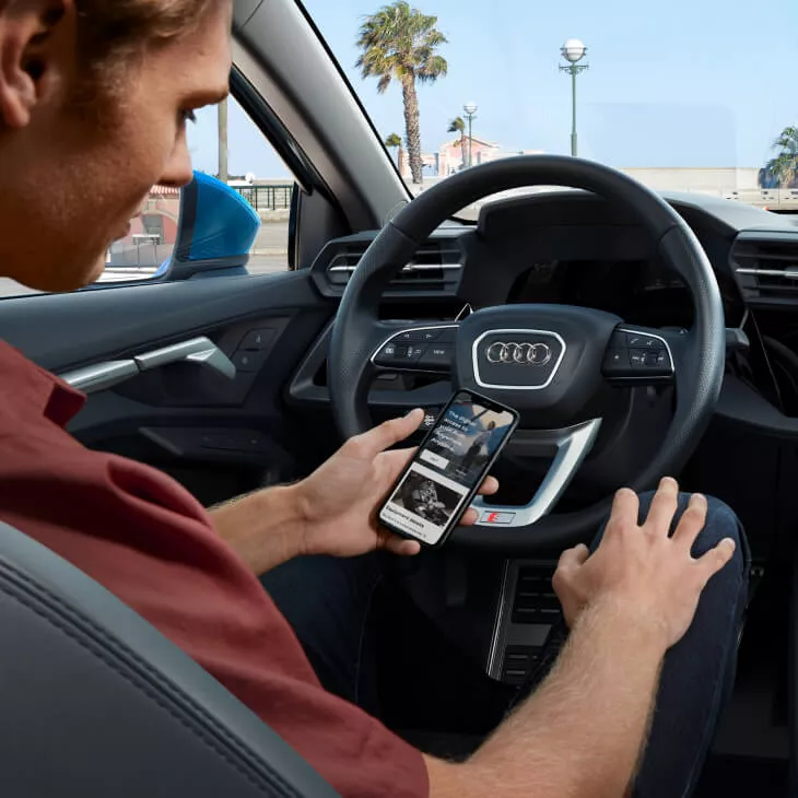 A male driver in sat in the driving seat of a park Audi vehicle looking at his mobile phone