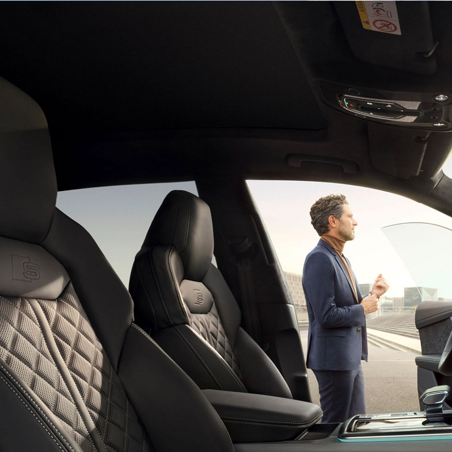 A suited man stands beside the open door on the driver side of an Audi vehicle