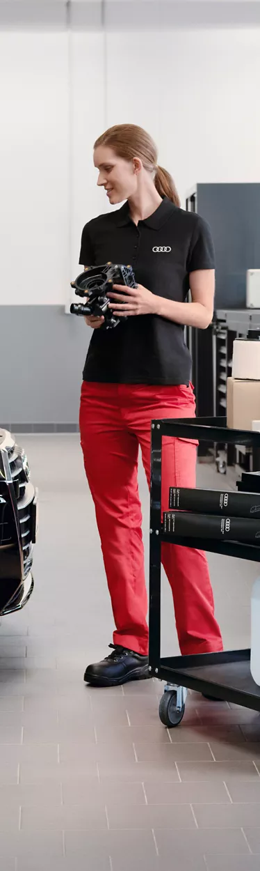 An Audi mechanic with an engine part in her hands looking at the engine the Audi vehicle beside her