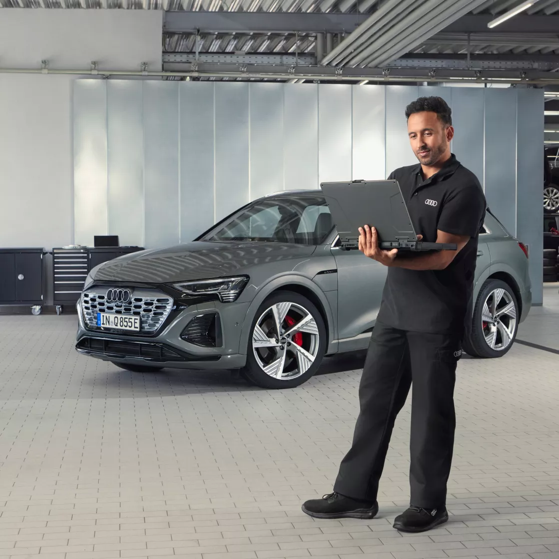 An Audi technician looking at a laptop with an Audi vehicle parked in the background