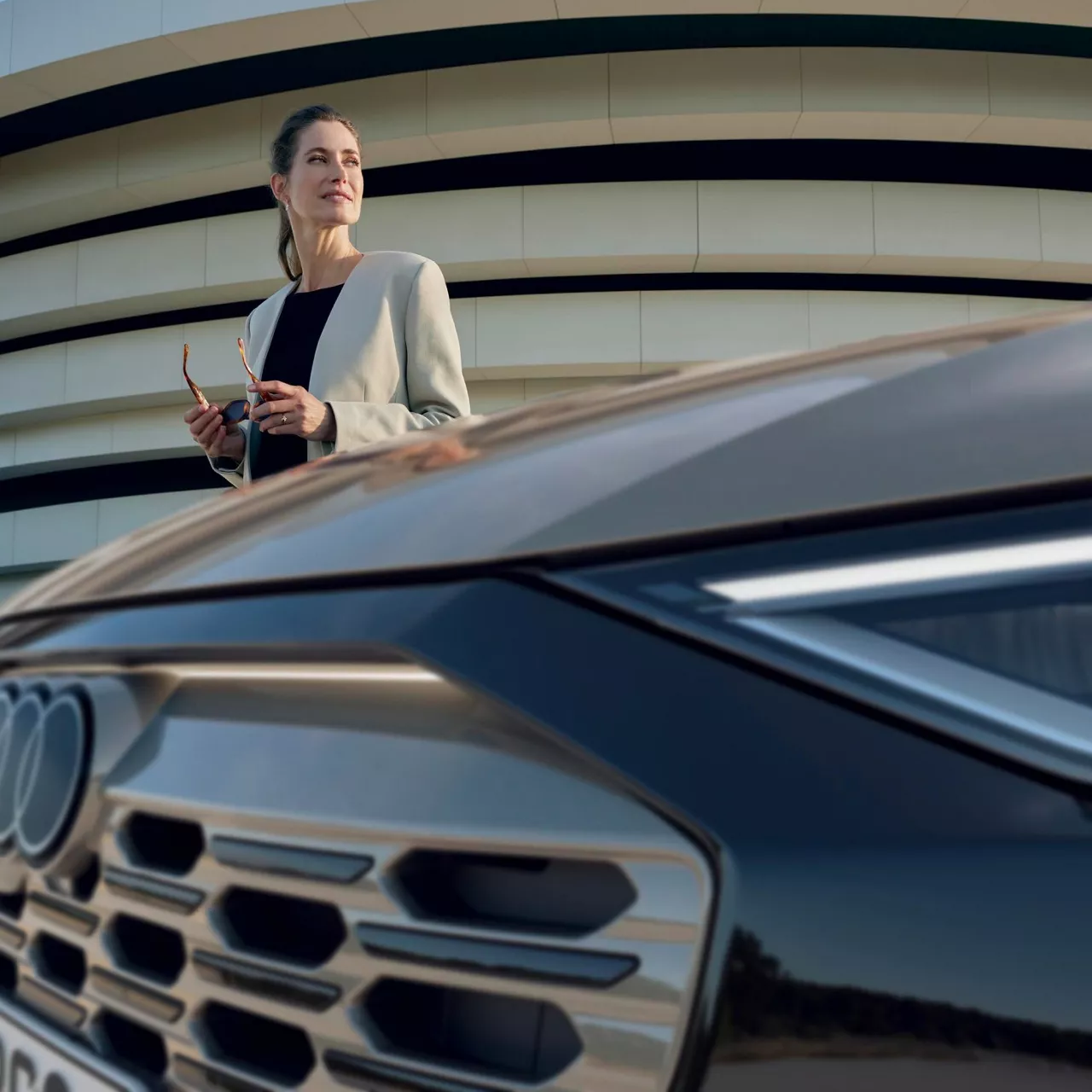 A woman with glasses in her hands standing beside the front of an Audi vehicle