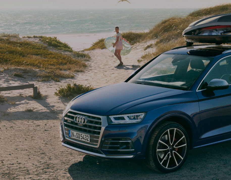 An Audi vehicle parked in a beach area with a young boy in the distance holding a child body board