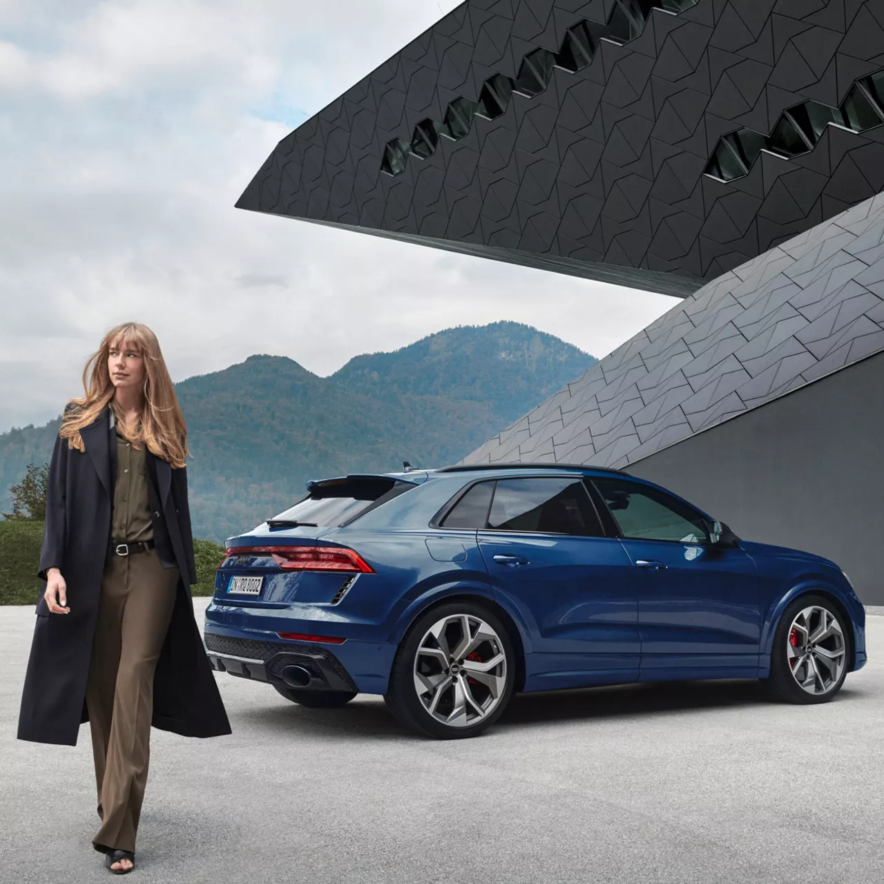 A young woman walking away from a blue parked Audi vehicle, modern house & mountains in the distance