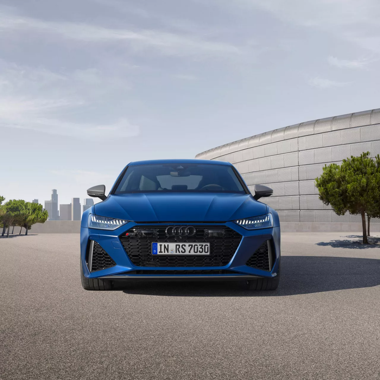 A blue Audi vehicle parked and facing us, trees and cityscape in the distance