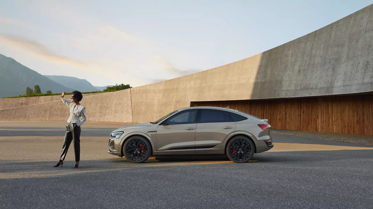 A person standing next to a parked Audi vehicle in a spacious environment in the late afternoon