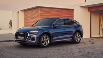 A blue electric Audi vehicle charging at a residential driveway during sunset