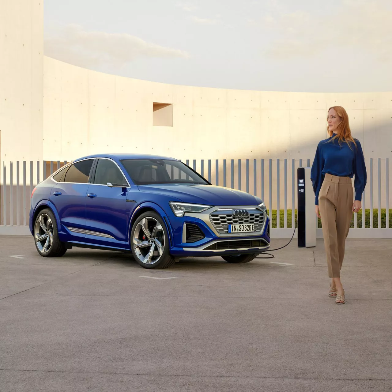 A woman walking away to the right of a parked blue Audi vehicle that is on charge