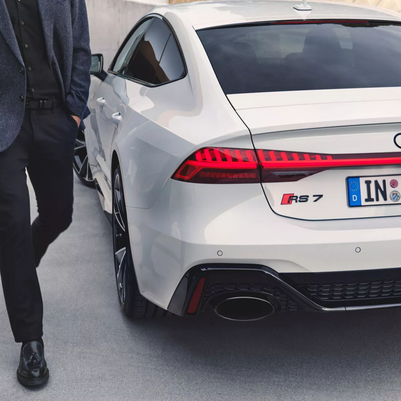 A suited man walking beside the rear left of a parked white Audi RS 7 vehicle