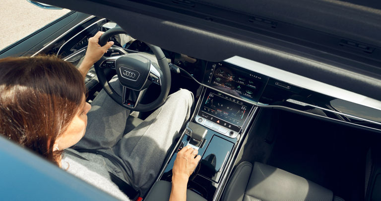 Person holding a steering wheel inside an Audi with a view of the dashboard.