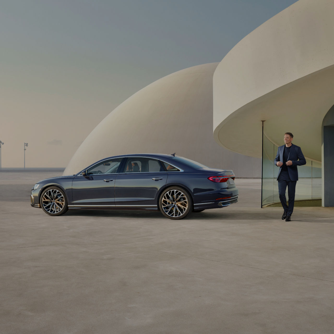 A sedan parked beside a modern building with a man in a suit walking towards it. 