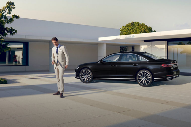 A person in a white suit walking towards a black car parked outside a modern building.