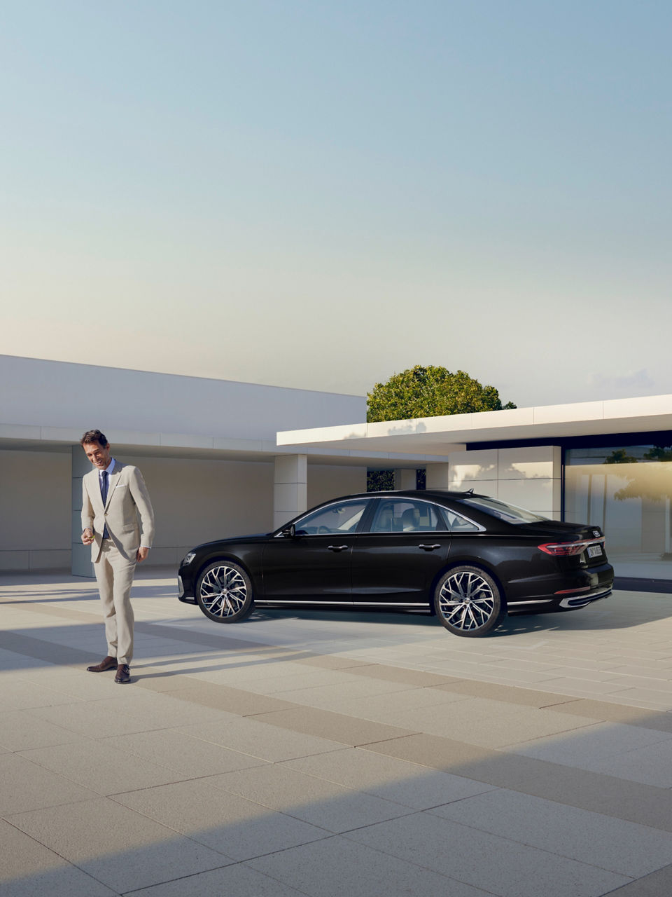 A person in a white suit walking towards a black car parked outside a modern building.