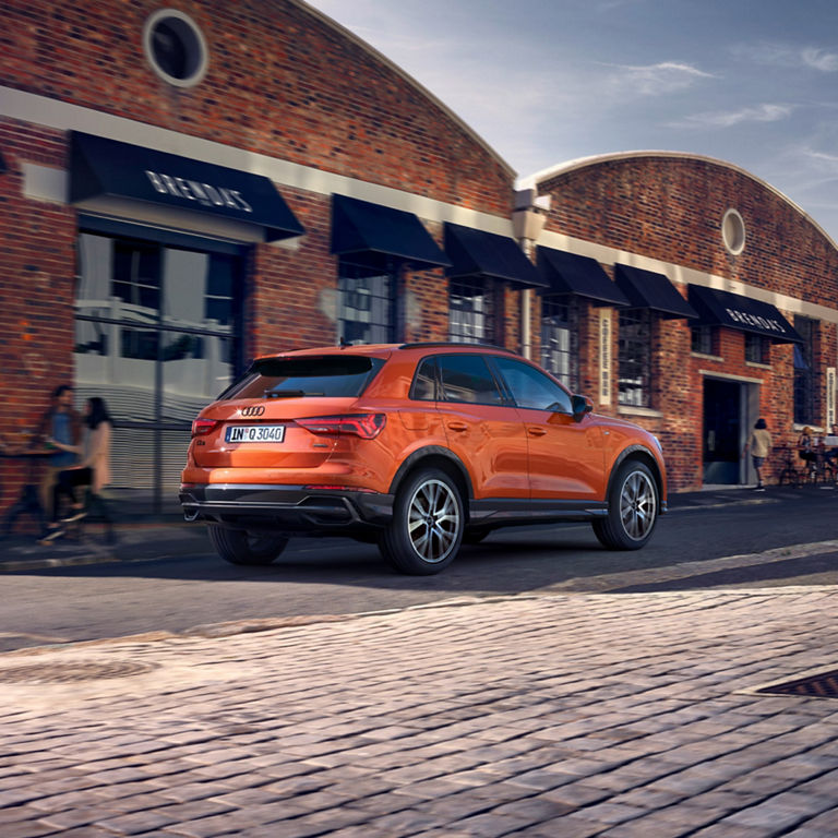 Perspective rear view of pulse orange Audi Q3 moving along residential street with  a blurred background.