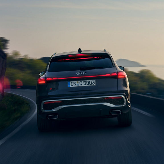 Audi SUV driving on a coastal road at dusk, with taillights on.