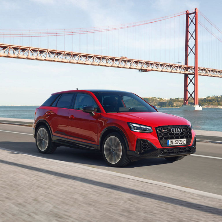 Red SUV driving on road by a large suspension bridge over water.