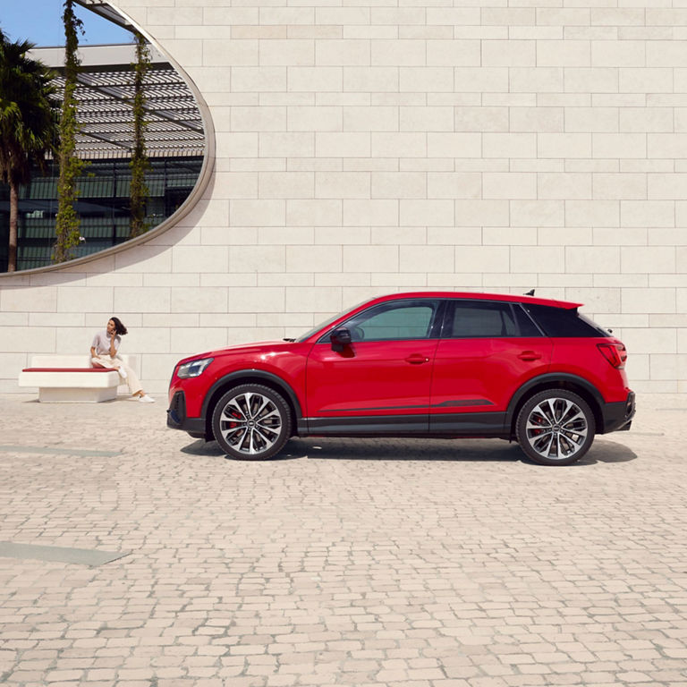 A red SUV parked beside a sitting area with a person on a bench outside a modern building.