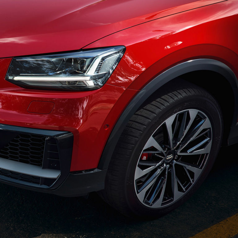 Closeup of red Audi car showing left side front grille, headlight and tyre. 